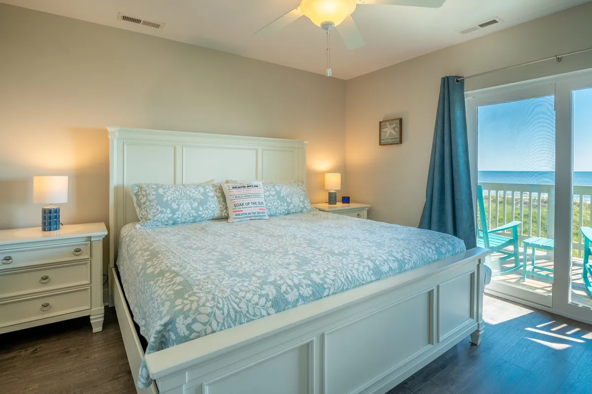 The image shows a spacious master bedroom featuring a large bed with floral bedding, nightstands with lamps on either side, and a door leading to a balcony. Soft natural light filters in through the balcony door, creating a calm and inviting atmosphere ideal for relaxation.