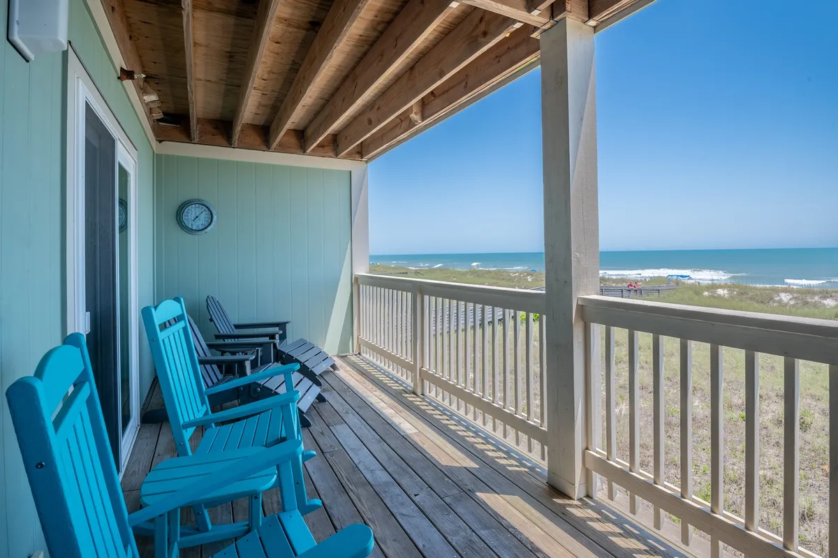 The hero image features a spacious wooden deck overlooking the ocean, with inviting blue rocking chairs perfectly positioned to enjoy panoramic sunrise views and the refreshing sea breeze. The tranquil setting suggests a perfect spot for sipping coffee, relaxing with a book, or sharing family moments against the backdrop of rolling waves.
