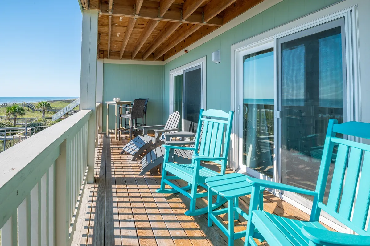 The image shows brightly colored Adirondack chairs and a pub table set on a wooden deck overlooking the ocean. The atmosphere feels cheerful and welcoming, inviting guests to enjoy breezy meals, morning coffee, or relaxed conversations while soaking in unobstructed coastal views.