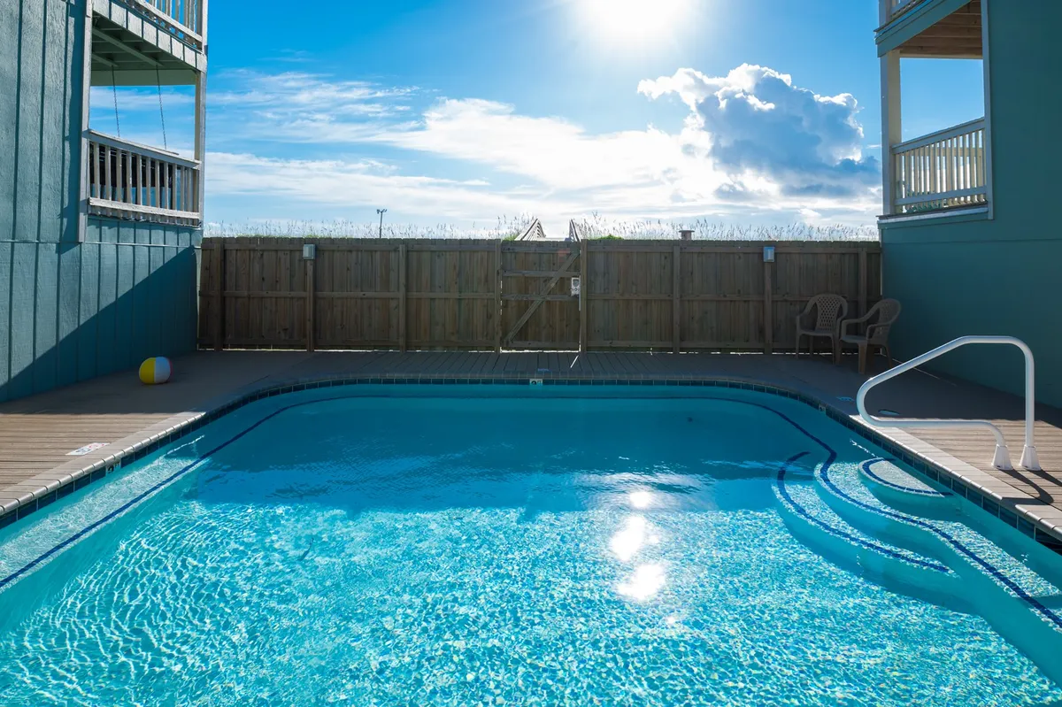 A bright, inviting swimming pool area catches sunlight, with a spacious patio and surrounding fence. The setting suggests relaxation, swimming, and opportunities for families to unwind together after beach adventures.