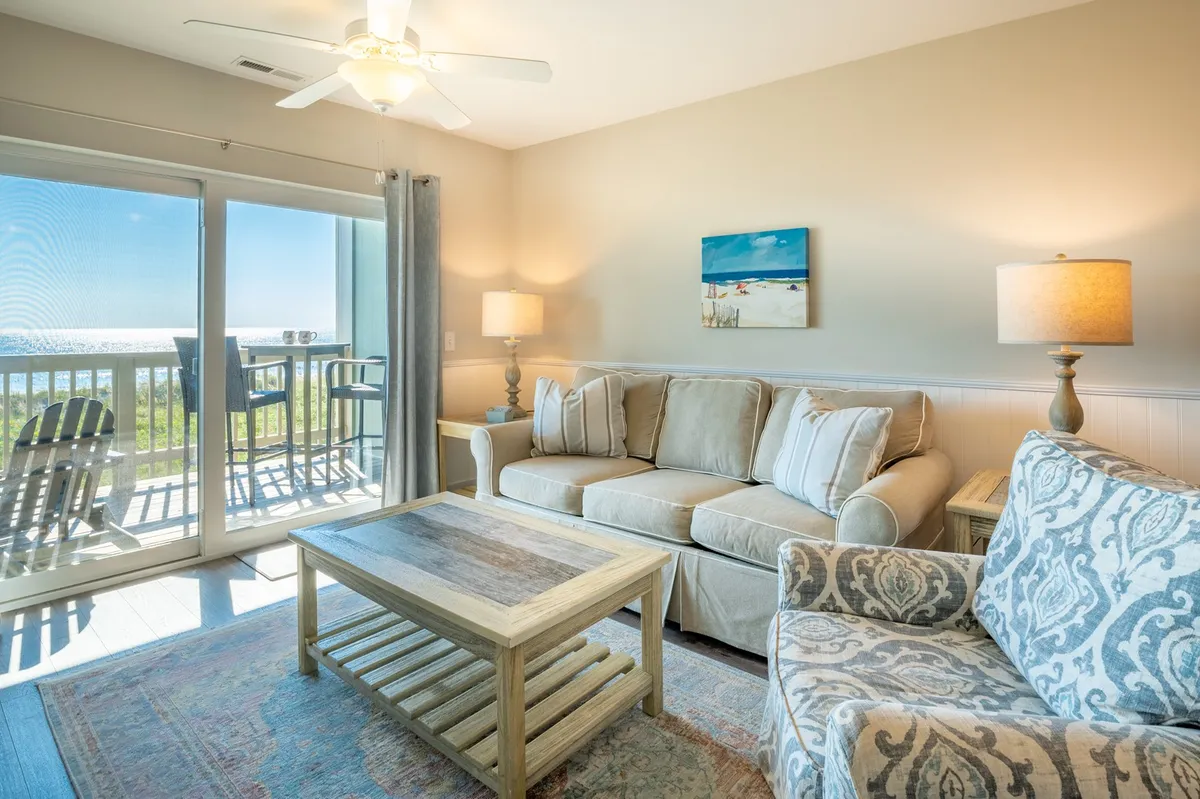 A cozy, inviting living area features a sofa and coffee table with a view of the outdoors. The space feels casual and perfect for socializing, with artwork sparking conversation and the balcony promising sunrise views—ideal for relaxing after a day out or preparing for a night on the boardwalk.