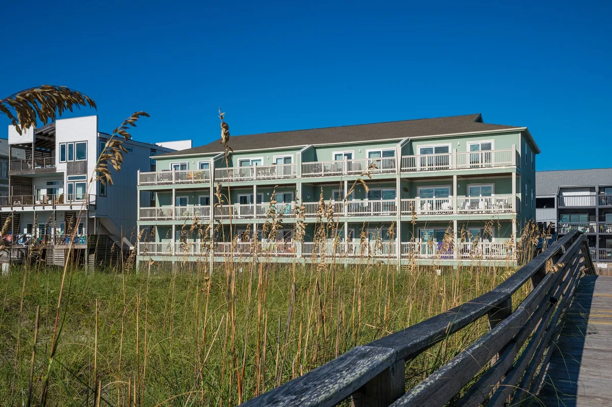 The exterior of the green condo building, with a welcoming wooden boardwalk stretching out over tall grasses and leading directly towards the beach. The sky is clear and blue, adding to the sense of openness and ease of access to the ocean.
