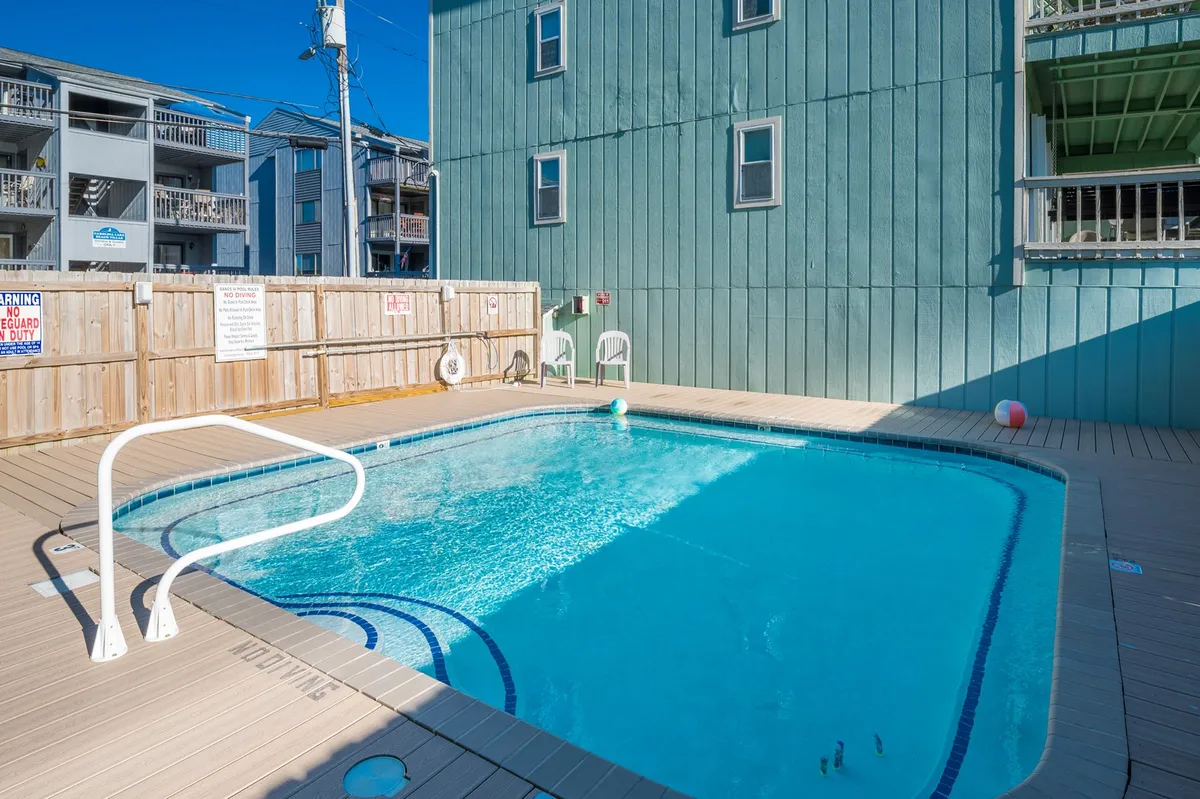An inviting swimming pool with a surrounding wooden deck adjacent to a green building facade. Guests are relaxing by the poolside, and the setting suggests a leisurely atmosphere perfect for both swimming and socializing.