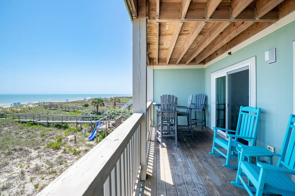 A private beachfront balcony furnished with comfortable seating overlooks the ocean. The wooden railings frame a sweeping view of the beach and surf, with the horizon visible in the distance. The tranquil setting suggests the perfect spot for morning coffee, sunset cocktails, and romantic moments while watching dolphins or the changing sky.