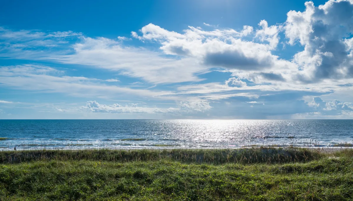 This image captures a sweeping view of the ocean surface, with clouds above and a grassy foreground, evoking the serene and inviting atmosphere of a classic beachfront location. The scene hints at opportunities for beachcombing, sunbathing, and picnicking just steps from the property.