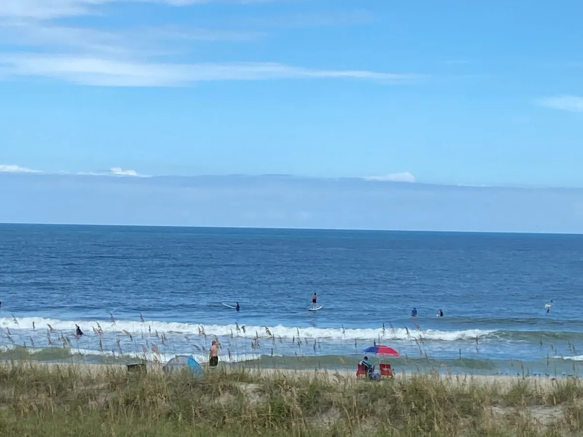 A lively scene of the beach with ocean waves, surfers, beachgoers, and families relaxing under umbrellas on provided beach chairs. The vibrant activity along the shore reflects a sense of fun and togetherness.