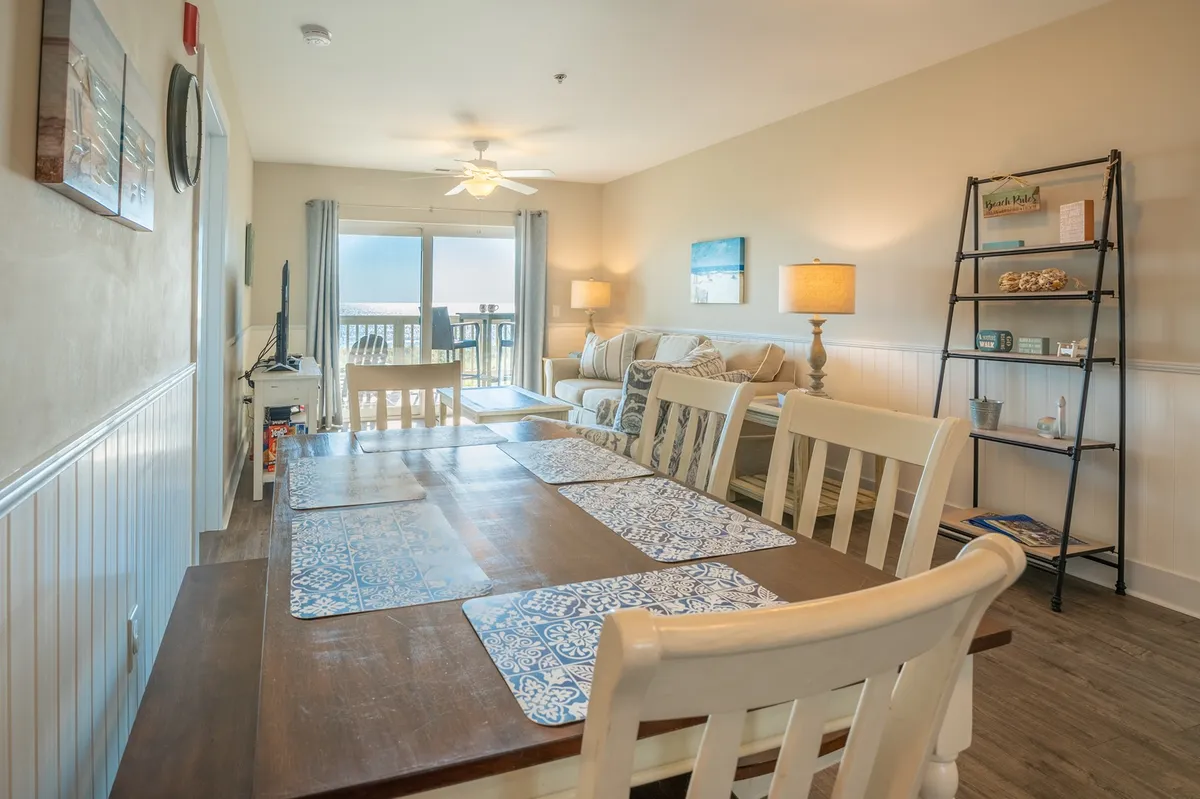 A welcoming, open-concept living and dining area featuring a comfortable sofa, spacious dining table, and large windows allowing natural light to pour in. The decorative shelf and window nook add warmth, while the layout invites socializing and family gatherings. Views from the balcony hint at the close connection to the oceanfront.