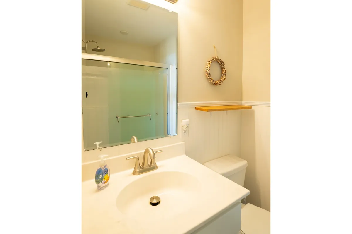 The image shows a bright bathroom with a modern sink, large mirror, a decorative wreath, and a glimpse of a walk-in shower. The setting feels clean, fresh, and inviting, offering a spa-like experience for personal care and relaxation.