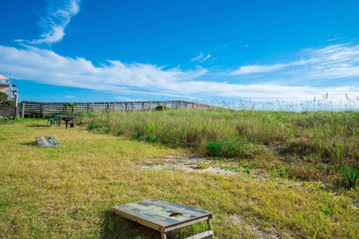 A casual outdoor space featuring cornhole boards on the grass, bordered by tall grass and a wooden railing. The atmosphere is laid-back, ideal for afternoon games under the sun.