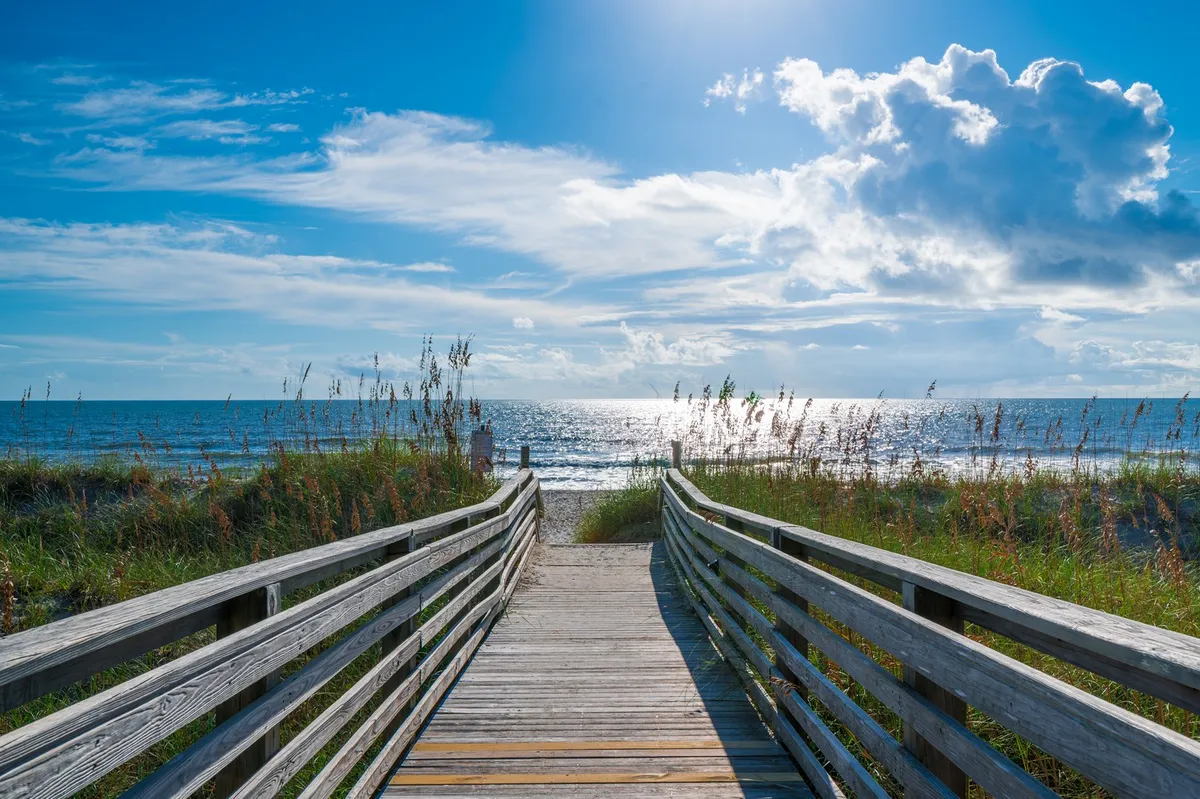 A tranquil wooden boardwalk stretches through dune grasses toward the ocean, with an expansive view of the sky and sea. The setting invites families to stroll to the beach or relax while taking in the scenery.